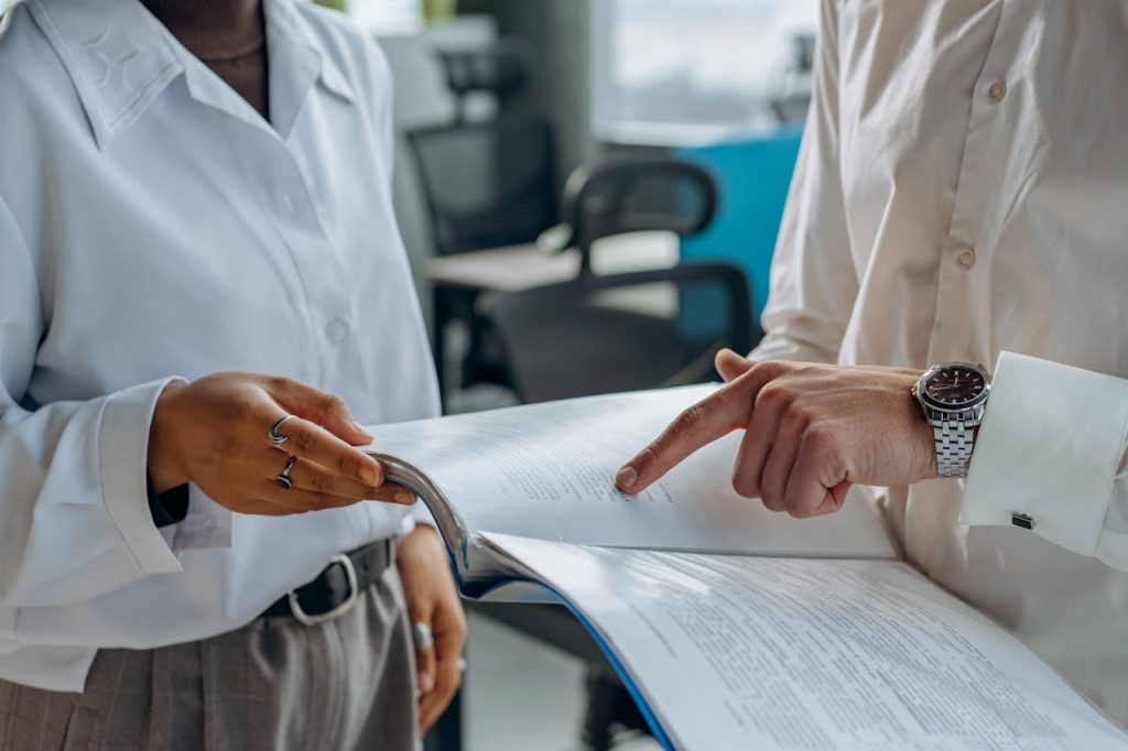 Two people in white shirts are reviewing a document together in an office setting. One person is holding the open document while the other points at a section on the pages. Both individuals are partially visible, focusing on the document.
