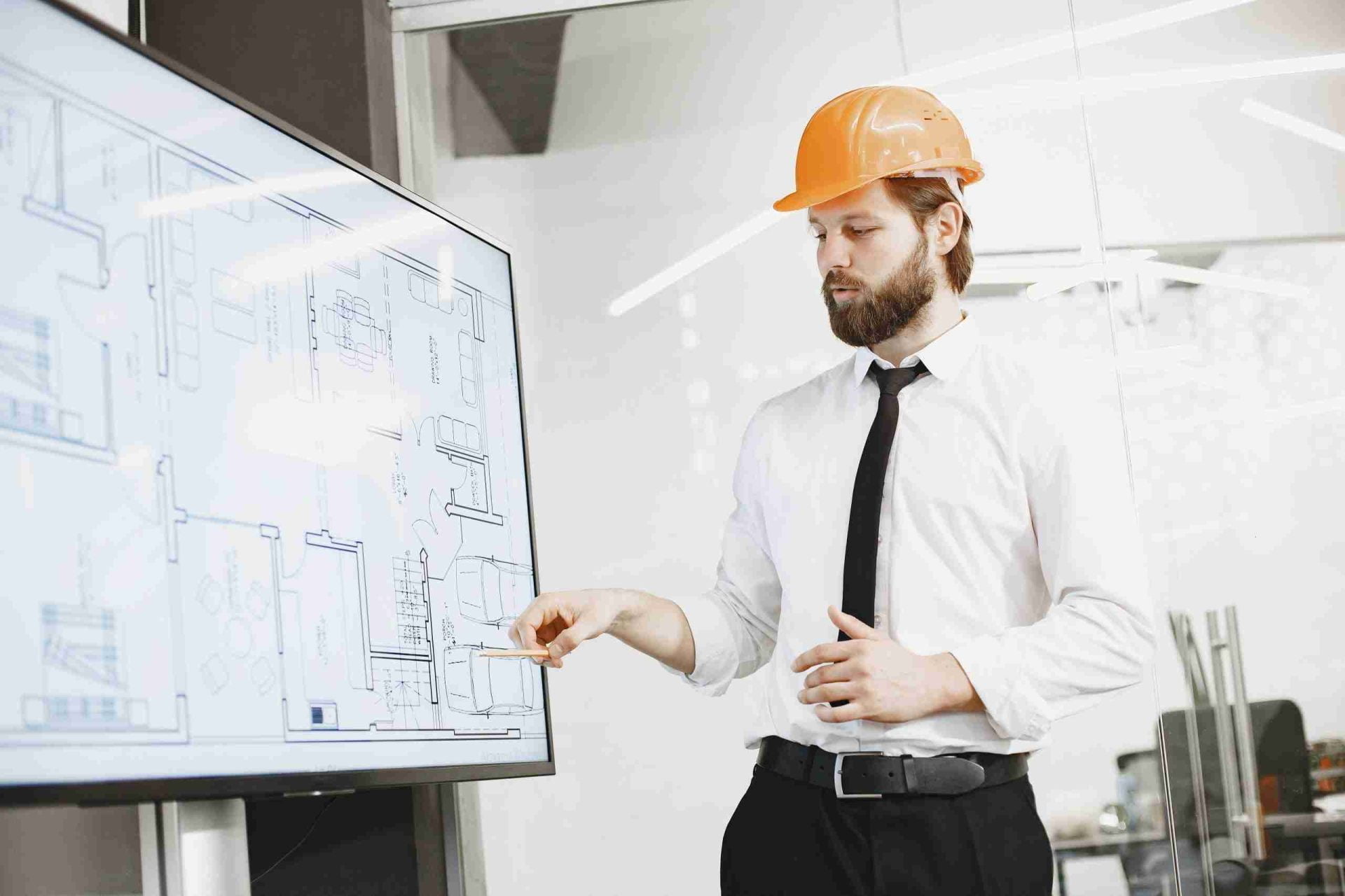 A man wearing an orange hard hat and a white shirt with a black tie points to a large screen displaying architectural plans. He is standing in a modern office space, explaining the details of the design.