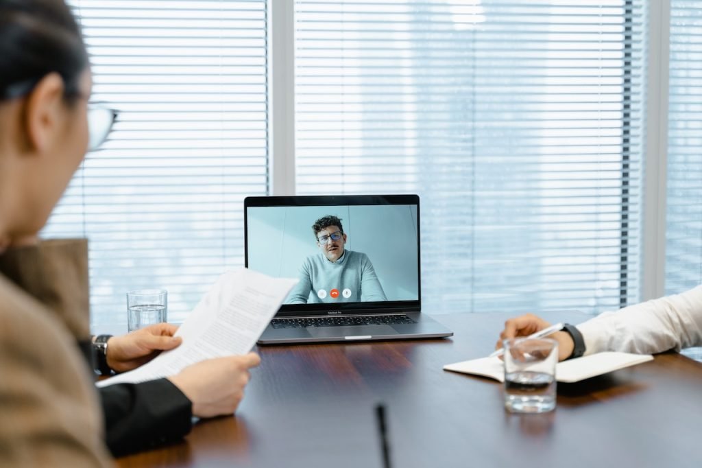 Two people sit at a conference table in front of a laptop during a video conference call. One person is holding a document, and the other is taking notes. A person on the screen is speaking and gesturing. There are glasses of water on the table.