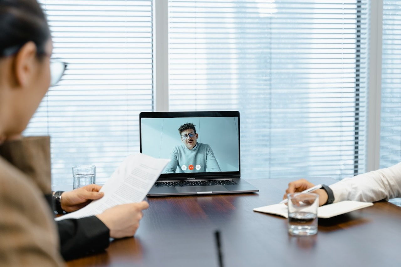 Two people sit at a conference table in front of a laptop during a video conference call. One person is holding a document, and the other is taking notes. A person on the screen is speaking and gesturing. There are glasses of water on the table.
