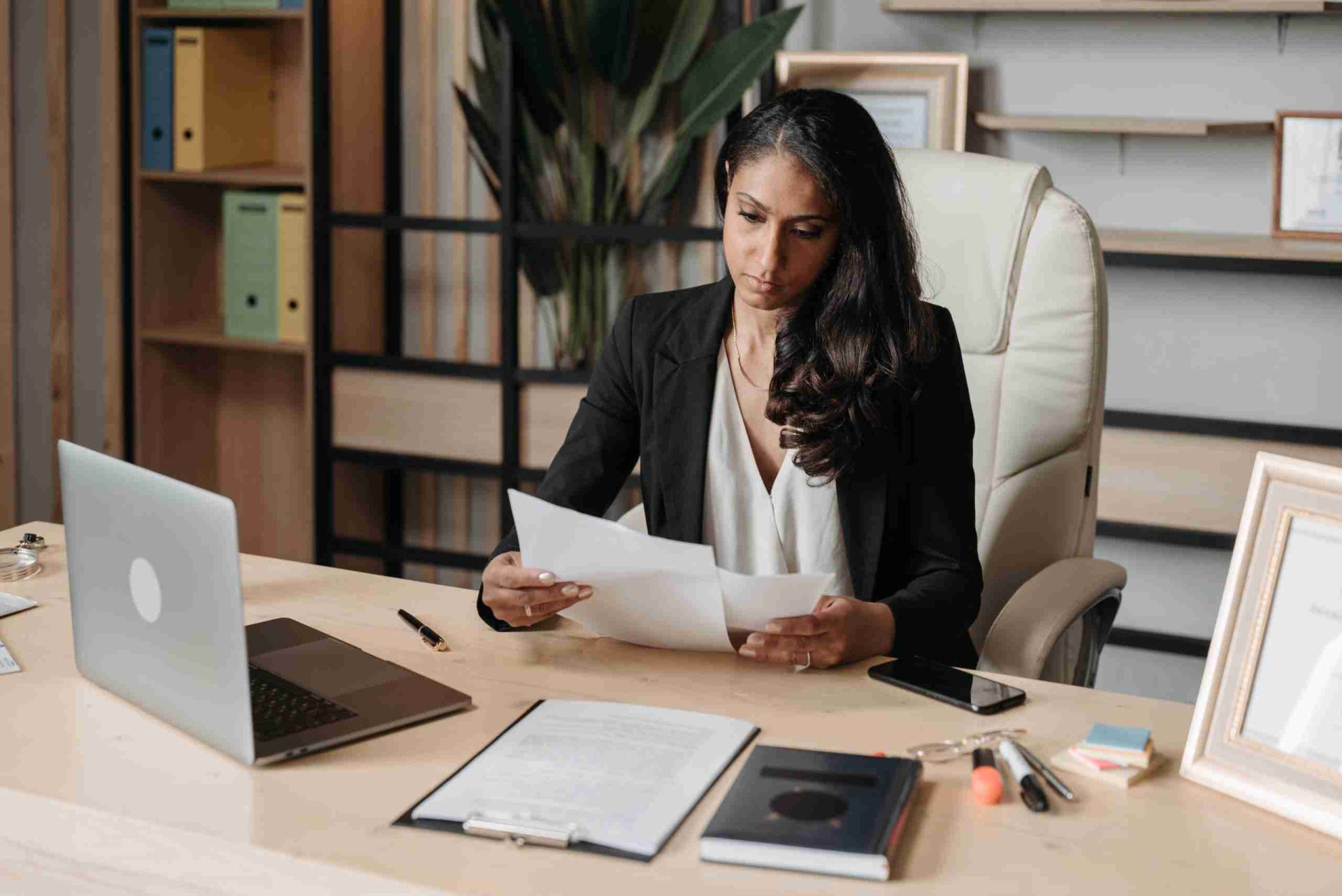 A woman in a black blazer is sitting at a desk in an office, examining documents. The desk is equipped with a laptop, clipboard, smartphone, and various office supplies. A large plant and shelves with binders are visible in the background.