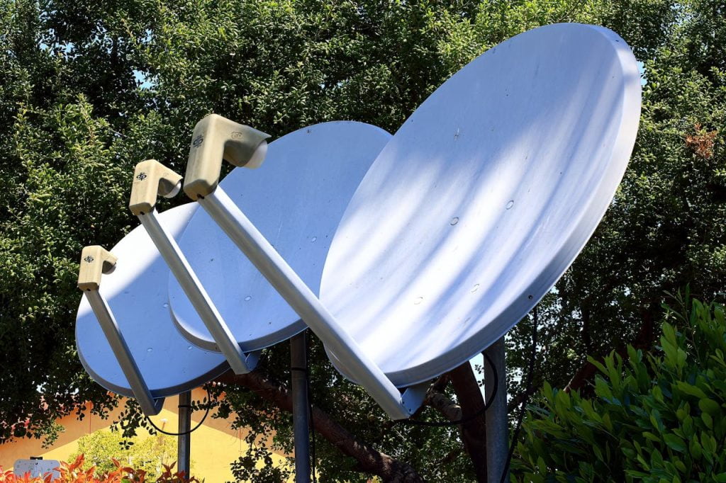Three large white satellite dishes are positioned outdoors, aimed skyward, with greenery and trees in the background. The dishes are mounted on sturdy poles and have metal support arms connecting to their receivers. The scene is bright and sunny.