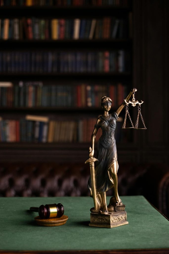 A bronze statue of Lady Justice holding scales and a sword stands on a green table. A wooden gavel and block sit nearby. The background features bookshelves filled with books, adding a legal and scholarly atmosphere to the scene.