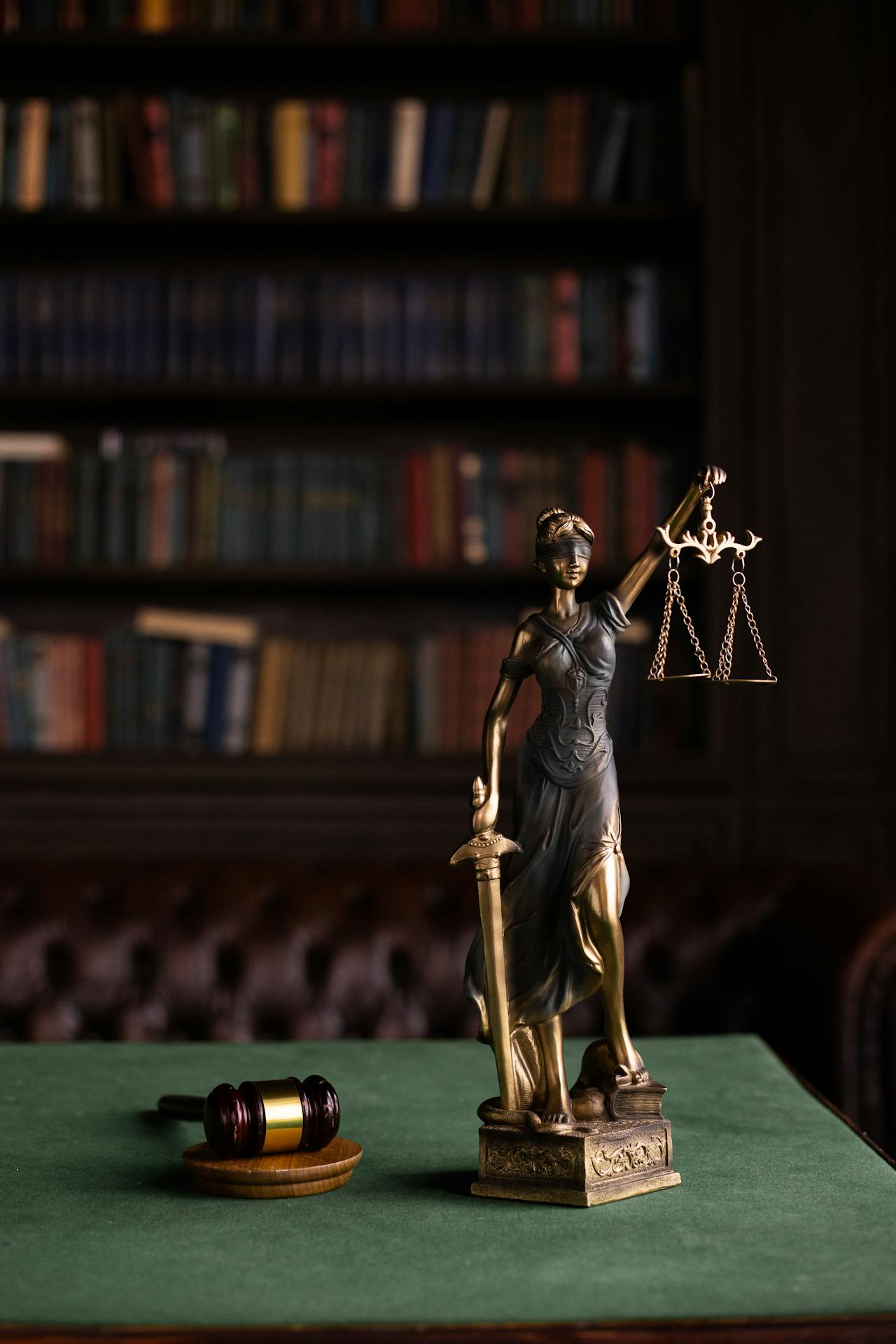 A bronze statue of Lady Justice holding scales and a sword stands on a green table. A wooden gavel and block sit nearby. The background features bookshelves filled with books, adding a legal and scholarly atmosphere to the scene.