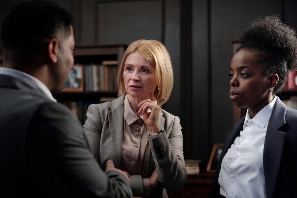 Three people in business attire are engaged in a discussion in an office. A woman with blonde hair is in the center, listening attentively with her hand on her chin. A man is on the left, and a woman with dark curly hair is on the right. Bookshelves are in the background.