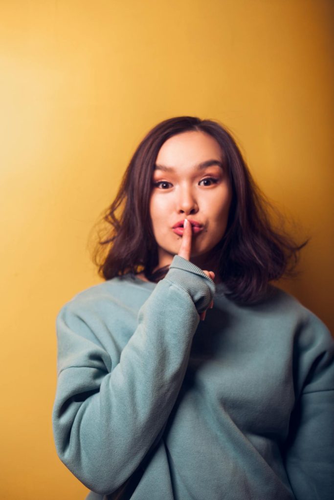 A woman with shoulder-length brown hair wearing a blue-green sweatshirt holds her index finger to her lips in a shushing gesture. She stands against a mustard yellow background.