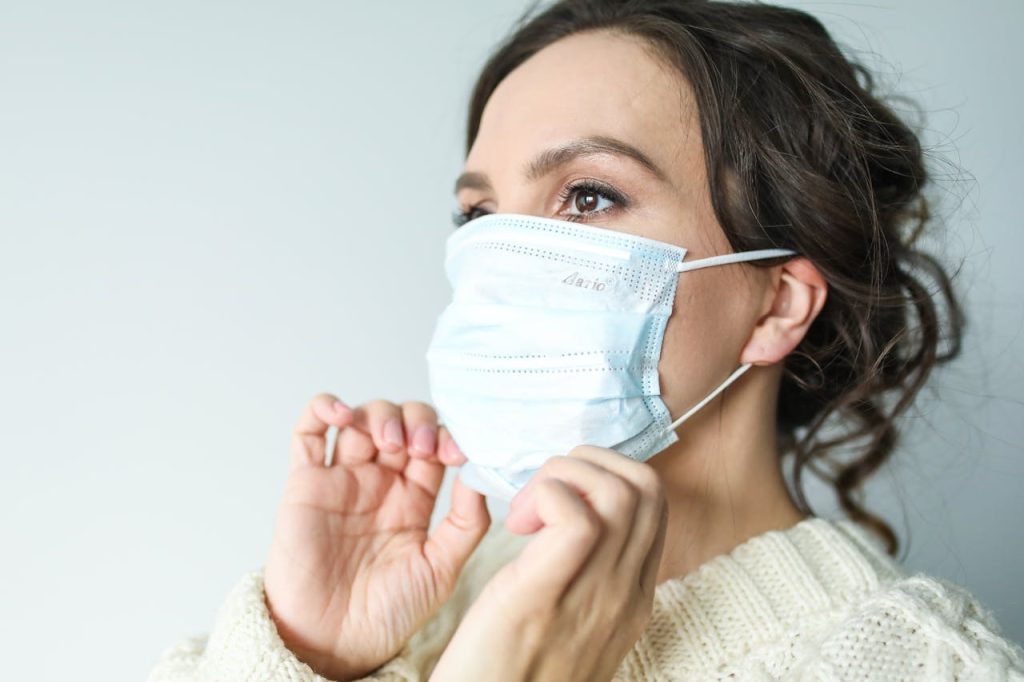 A woman with curly hair is wearing a light-colored sweater and adjusting a blue surgical mask on her face. The background is plain and light-colored.