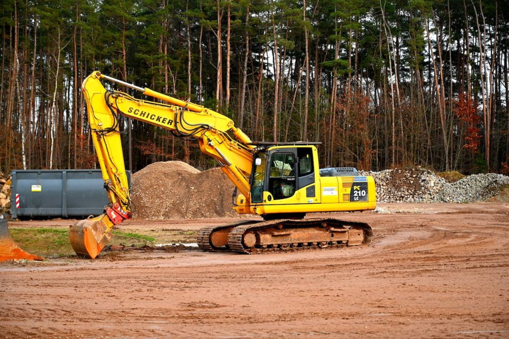 A yellow excavator with "Schicker" written on the arm is parked on a dirt clearing at a construction site, surrounded by piles of gravel and dirt. A forested area is visible in the background, with a large grey container to the left.