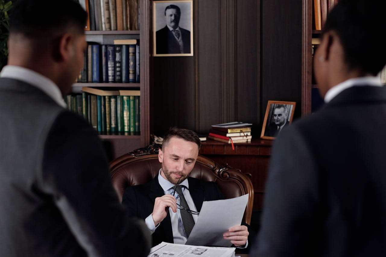 A man sitting at a desk in an office room holds a document and a pen, engaged in conversation with two standing individuals in business attire. The room features bookshelves filled with books and framed portraits on the wall in the background.