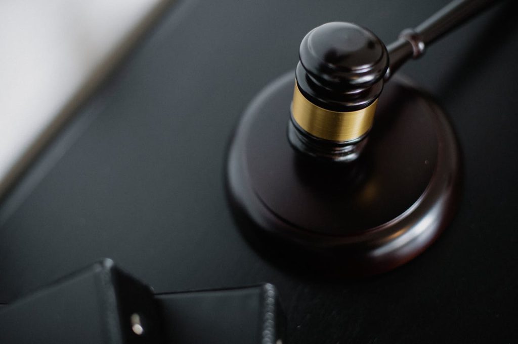 A close-up of a judge's gavel resting on a sound block, placed on a dark surface. The gavel, made of dark wood, features a metallic gold band around its head. A wallet or similar item lies slightly blurred in the foreground.
