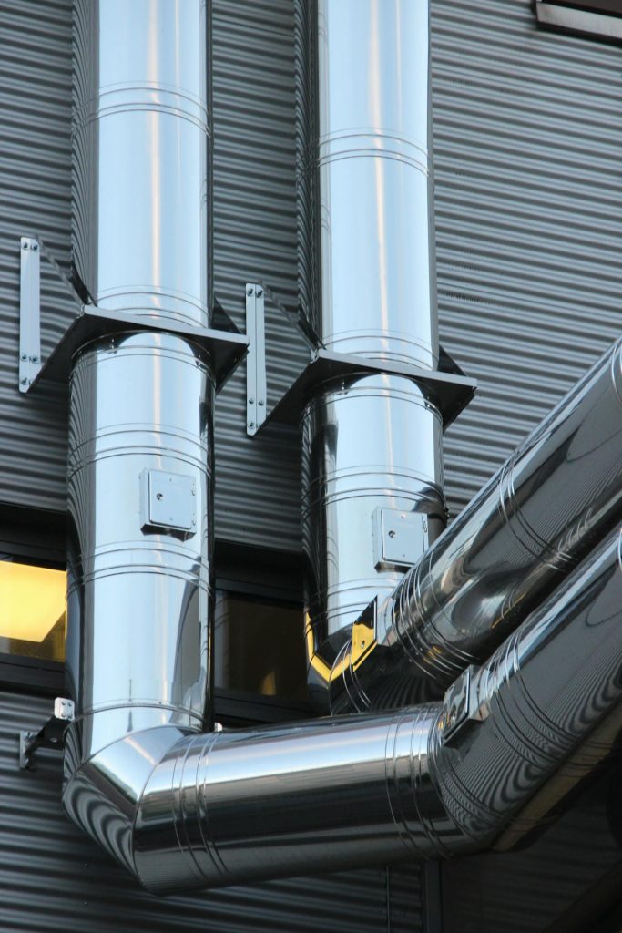 Close-up of shiny metal industrial pipes against a ribbed gray wall. The pipes, reflecting the surroundings, are connected at right angles and fitted with brackets to hold them in place. A small window with a yellow light can be seen partially behind the pipes.