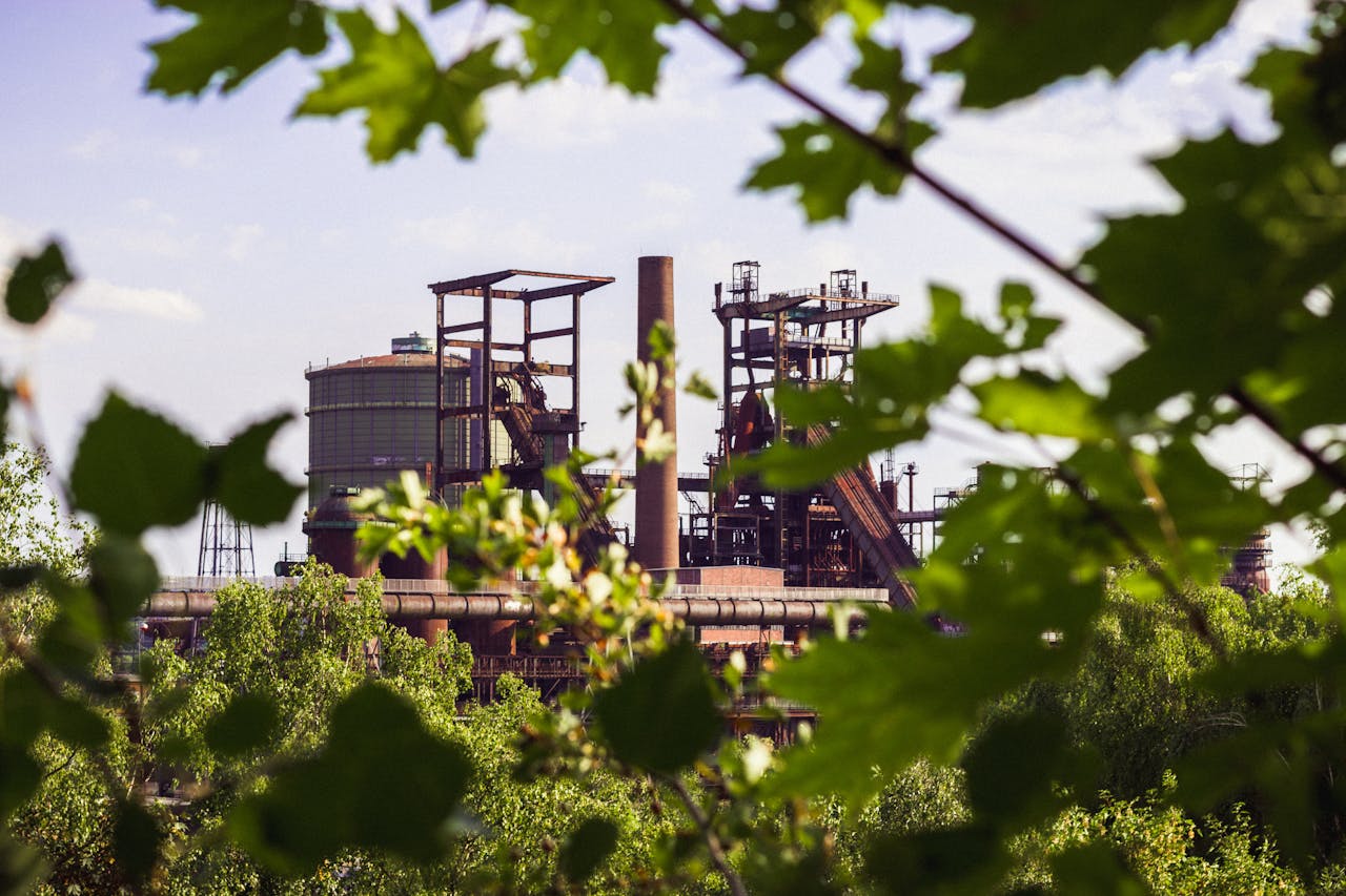 A distant view of an industrial facility with tall structures, cylindrical tanks, and smokestacks, partially obscured by lush green leaves and branches in the foreground. The sky is bright with scattered clouds.