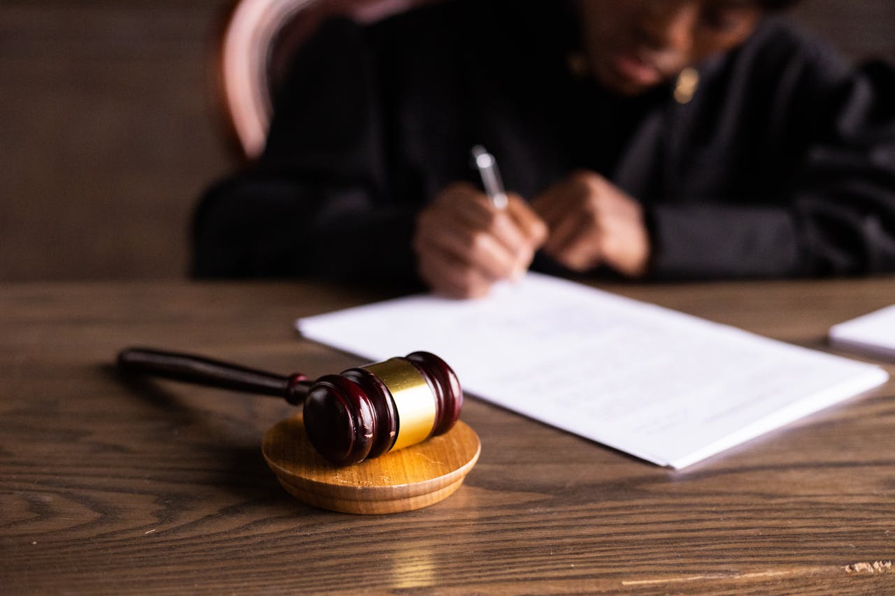 A judge's gavel rests on a wooden desk in the foreground, symbolizing justice. In the background, slightly out of focus, a person in a judicial robe writes on a piece of paper, possibly signing legal documents or taking notes.