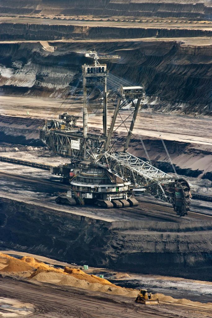 A massive industrial excavator, likely used in mining, with multiple large mechanical arms and a rotating bucket-wheel, is operating in a vast open-pit mine. The earth around it is tiered in layers, showing extensive excavation work.
