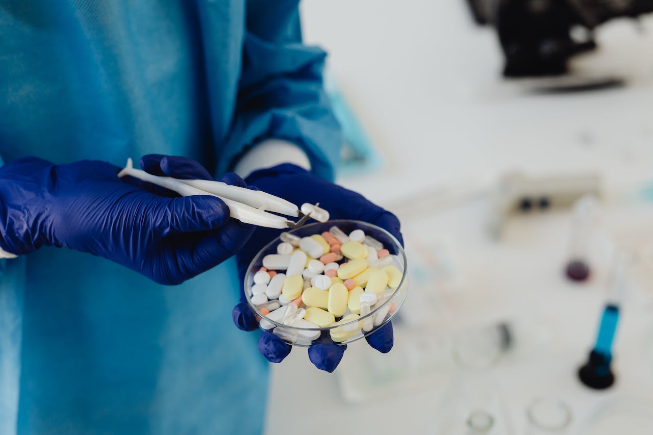 A person in a blue lab coat and blue gloves uses tweezers to pick up a pill from a petri dish filled with various colorful pills. The background is a laboratory setting with scientific equipment.
