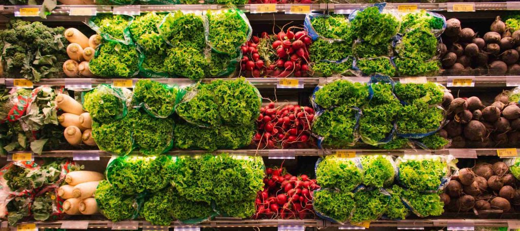 Grocery store produce section with shelves filled with fresh greens, including bags of lettuce and bunches of radishes. The vibrant green and red vegetables are neatly arranged in rows under bright lighting.