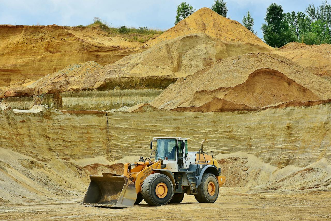 A large yellow and white front loader with a raised scoop is parked in a sand quarry. Behind the loader are various layers of sand and soil in different shades, and in the distance, there are green trees atop the sandy hills.