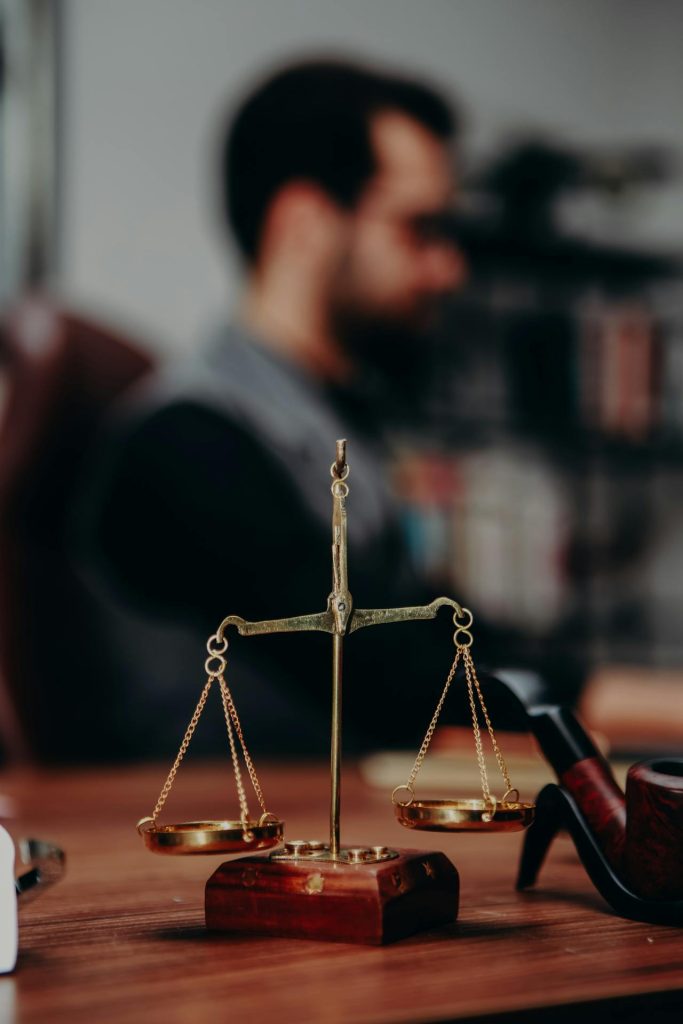 A brass balance scale sits prominently on a wooden desk, with a pipe beside it. In the background, a blurred figure is seated, suggesting a setting in an office or study. The focus remains on the symbolic scales of justice.
