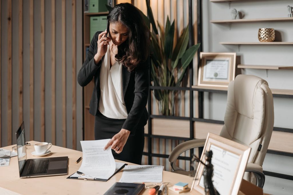 A woman stands at an office desk, talking on the phone while pointing at a document. The desk is cluttered with papers, a laptop, a cup of coffee, and various office supplies. Shelves with books, framed certificates, and decorations are in the background.