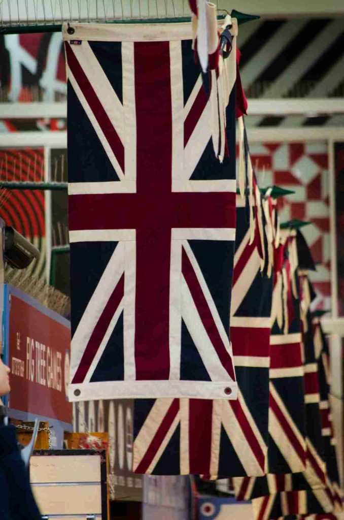 A row of Union Jack flags hanging vertically in a market or indoor space. The flags are prominently displayed, with the closest flag in clear focus, and others receding into the background. The scene includes various patterns and merchandise on display.