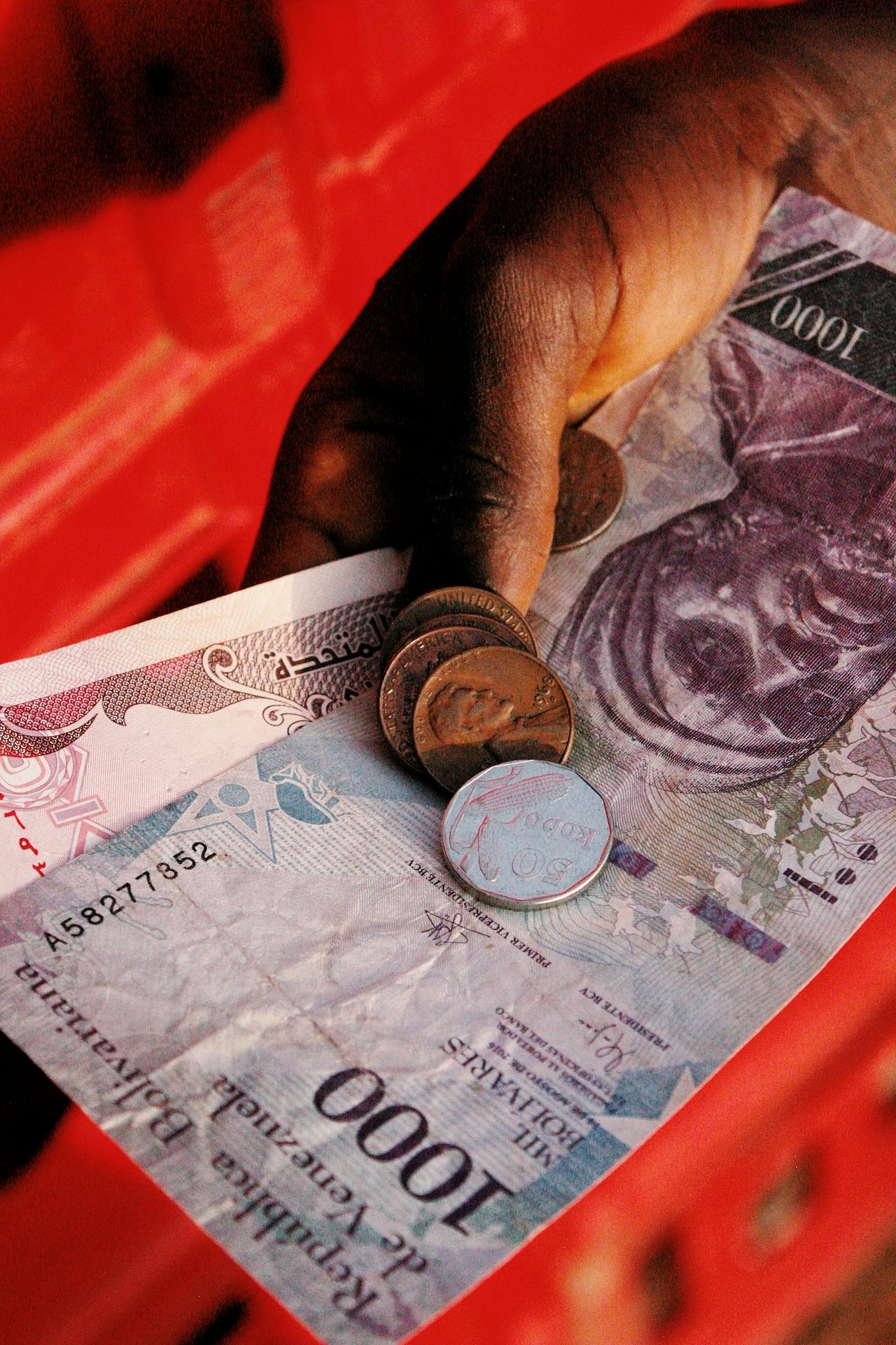 A hand holding Nigerian currency, including a 1000 Naira note and several coins, against a red background. The hand is slightly curled around the money, and the coins are stacked on top of the note.
