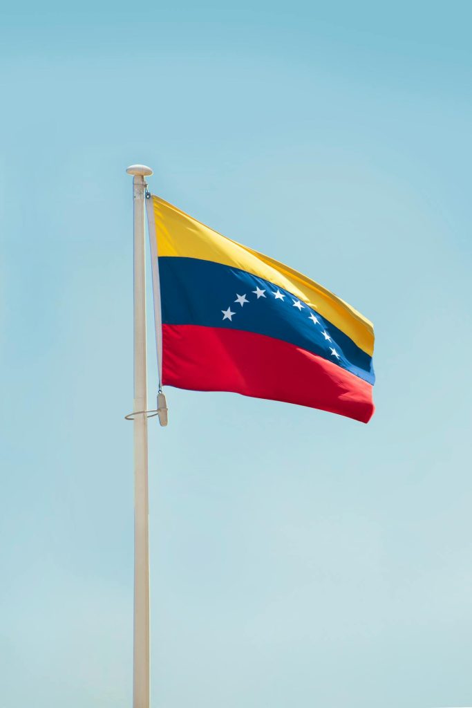 The image displays the flag of Venezuela waving on a flagpole against a clear blue sky. The flag consists of three horizontal stripes: yellow on top, blue in the middle with eight white stars in an arc, and red at the bottom.
