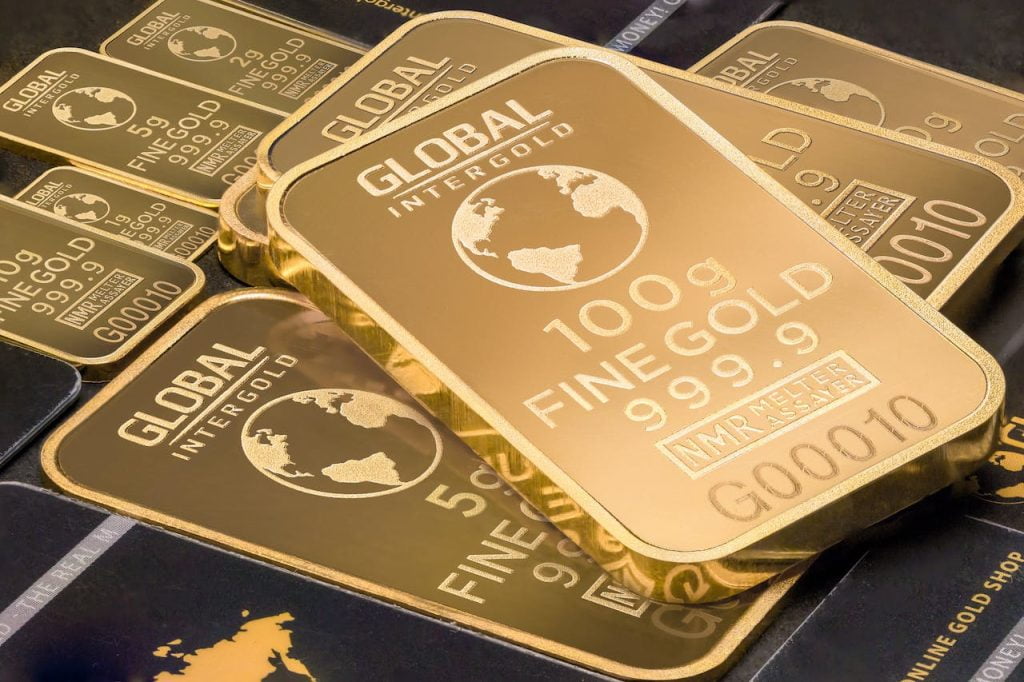 A close-up image of several gold bars of varying sizes. The largest bar prominently displays "GLOBAL INTERGOLD," "100 g," "FINE GOLD," "999.9," and other specifications. Smaller bars with similar inscriptions are scattered around it.