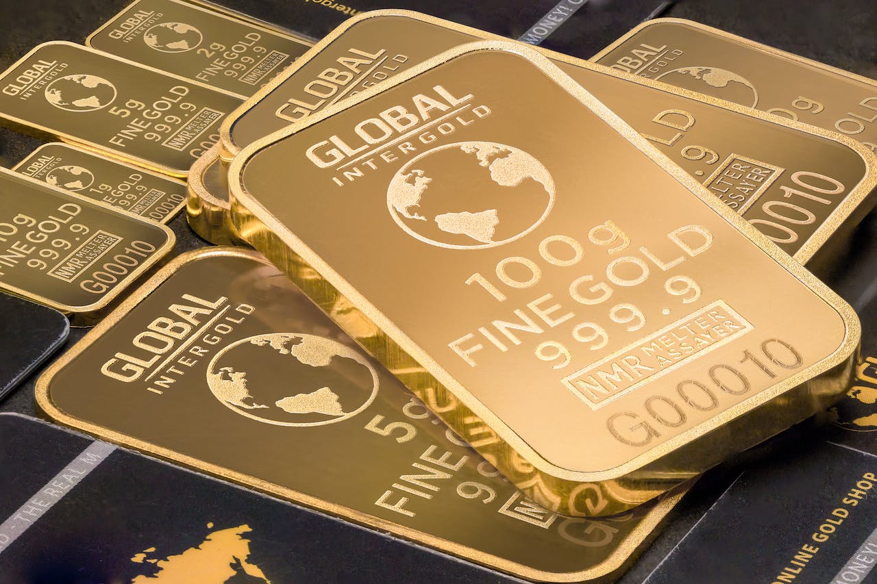 A close-up image of several gold bars of varying sizes. The largest bar prominently displays "GLOBAL INTERGOLD," "100 g," "FINE GOLD," "999.9," and other specifications. Smaller bars with similar inscriptions are scattered around it.