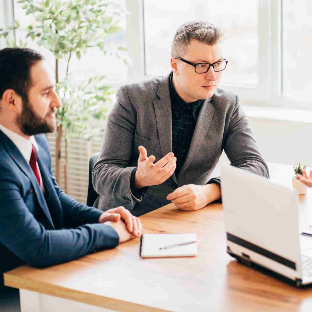 Two men in business attire sit at a wooden desk during a meeting. One, wearing glasses and a gray blazer, gestures while speaking. The other, in a blue suit and red tie, listens attentively. A notebook and laptop are on the desk, and a potted plant is in the background.