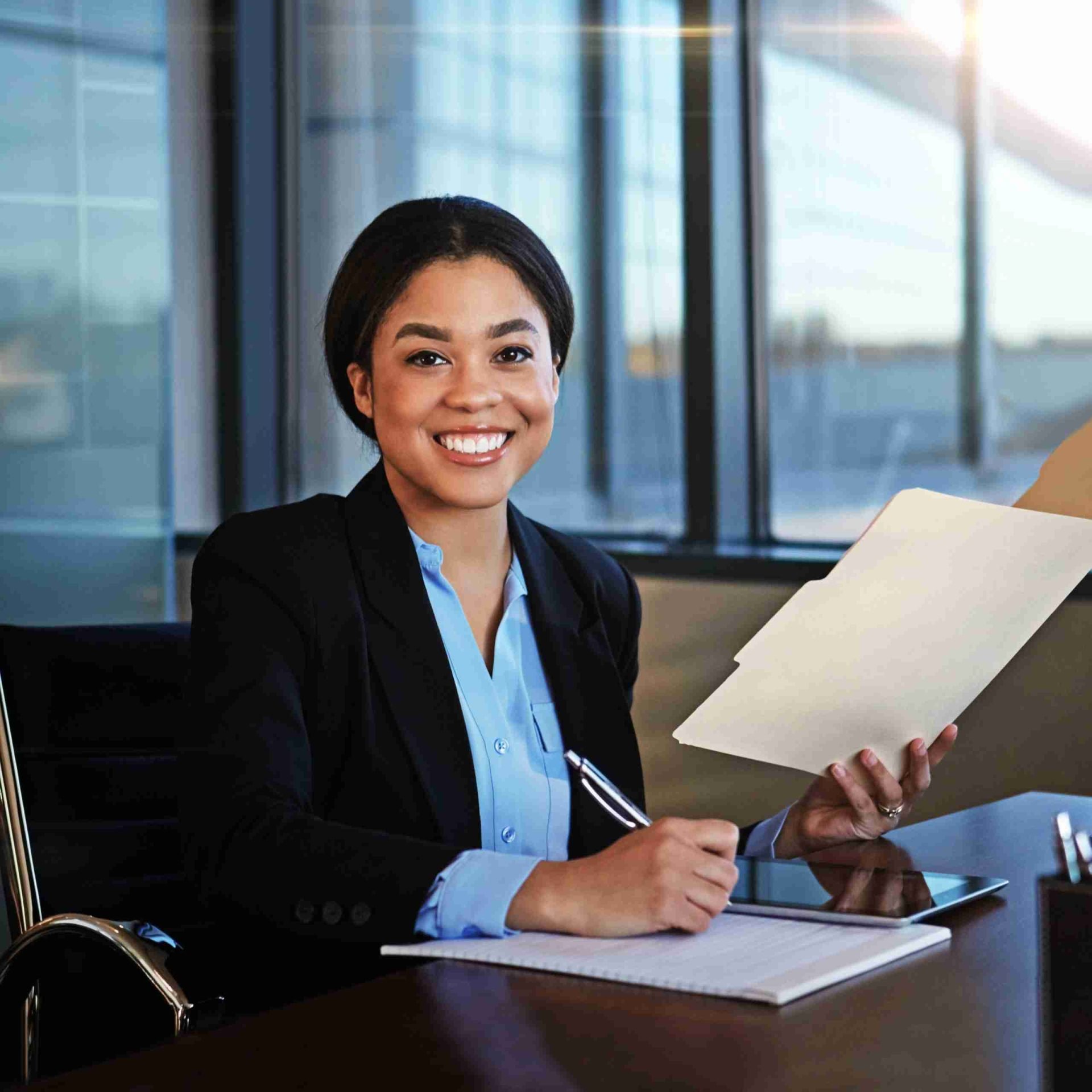 A young professional woman sits at a desk in a modern office. She is smiling, holding a pen in her right hand, and a folder with paperwork in her left hand. She is dressed in business attire, with a blazer and blue shirt. Large windows with a city view are behind her.
