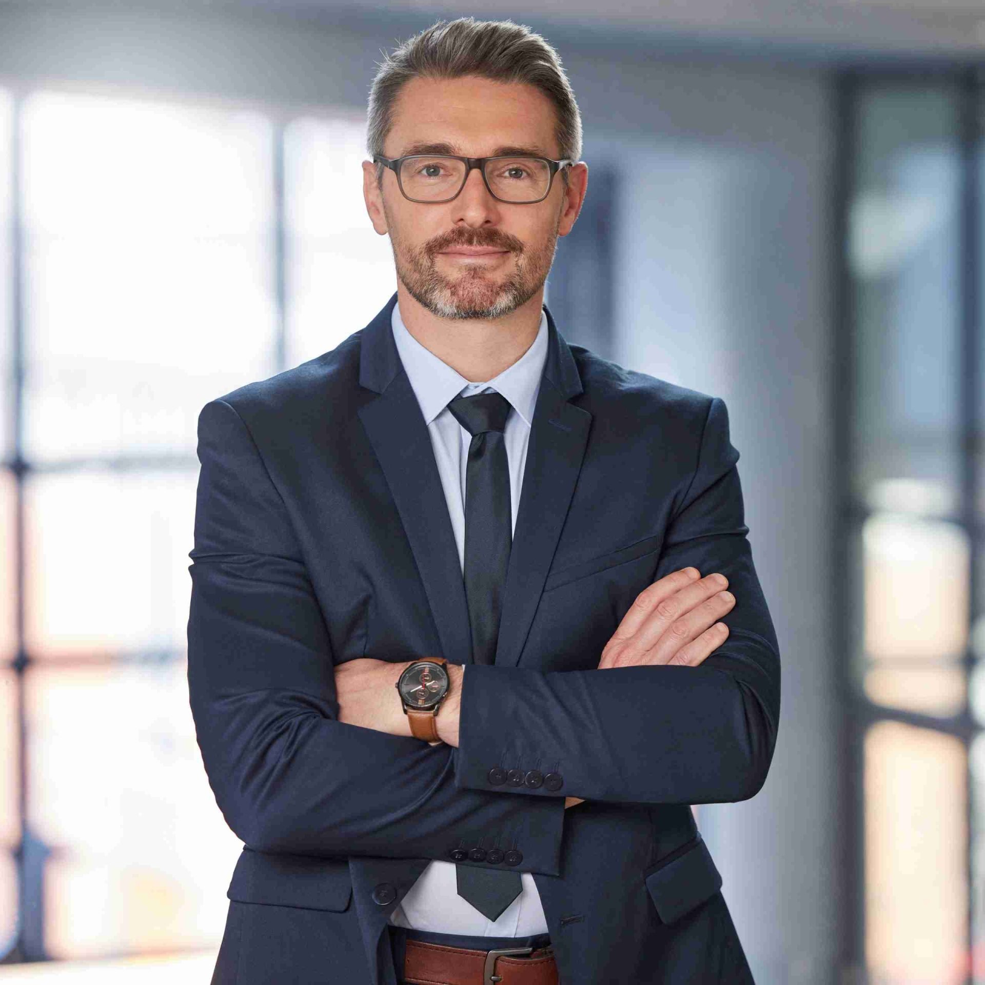 A man with short hair and glasses is standing with his arms crossed, wearing a dark suit, white dress shirt, and tie. He has a wristwatch on his left wrist. The background is blurred, suggesting an office setting with large windows.