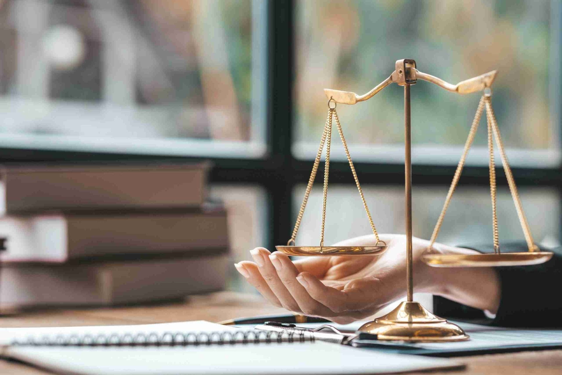 A close-up of a person's hand resting on a desk, with a set of golden scales in the foreground. They are in front of a window. There are stacks of books and a spiral notebook on the desk. The scales symbolize balance and justice.