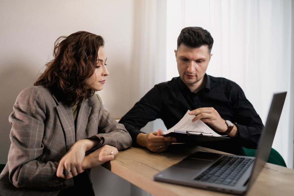 A man and woman sit at a wooden table with a laptop in front of them. The man, wearing a black shirt, holds a clipboard and explains something to the woman, who wears a gray blazer. The room has white walls and a curtain in the background.