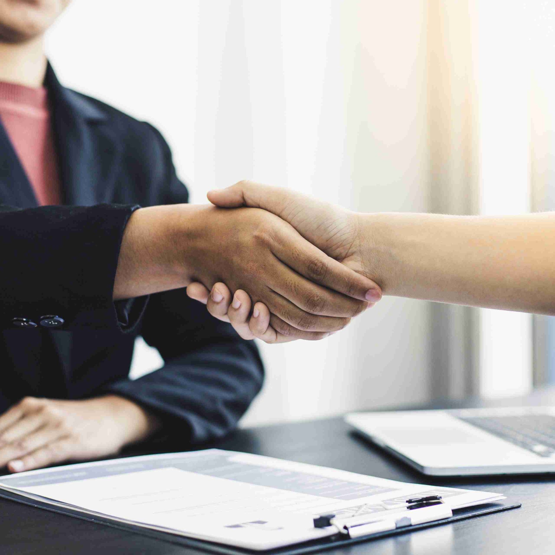 Two people shaking hands across a desk, one wearing a dark suit and the other without a visible outfit. A document and a pen are on the desk, with a laptop partially visible beside them, suggesting a business or professional agreement.