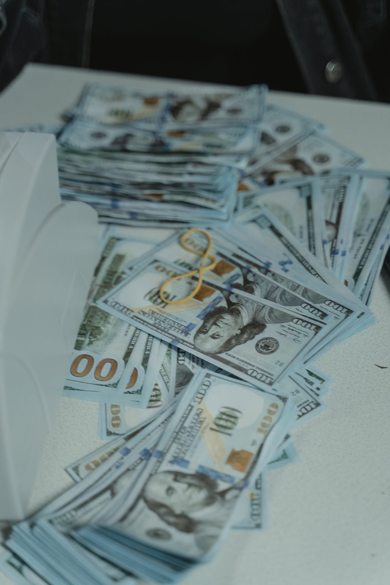 A scattered pile of U.S. hundred-dollar bills is spread across a white surface. A pair of yellow rectangular glasses rests on top of the pile, adding a splash of color to the scene.