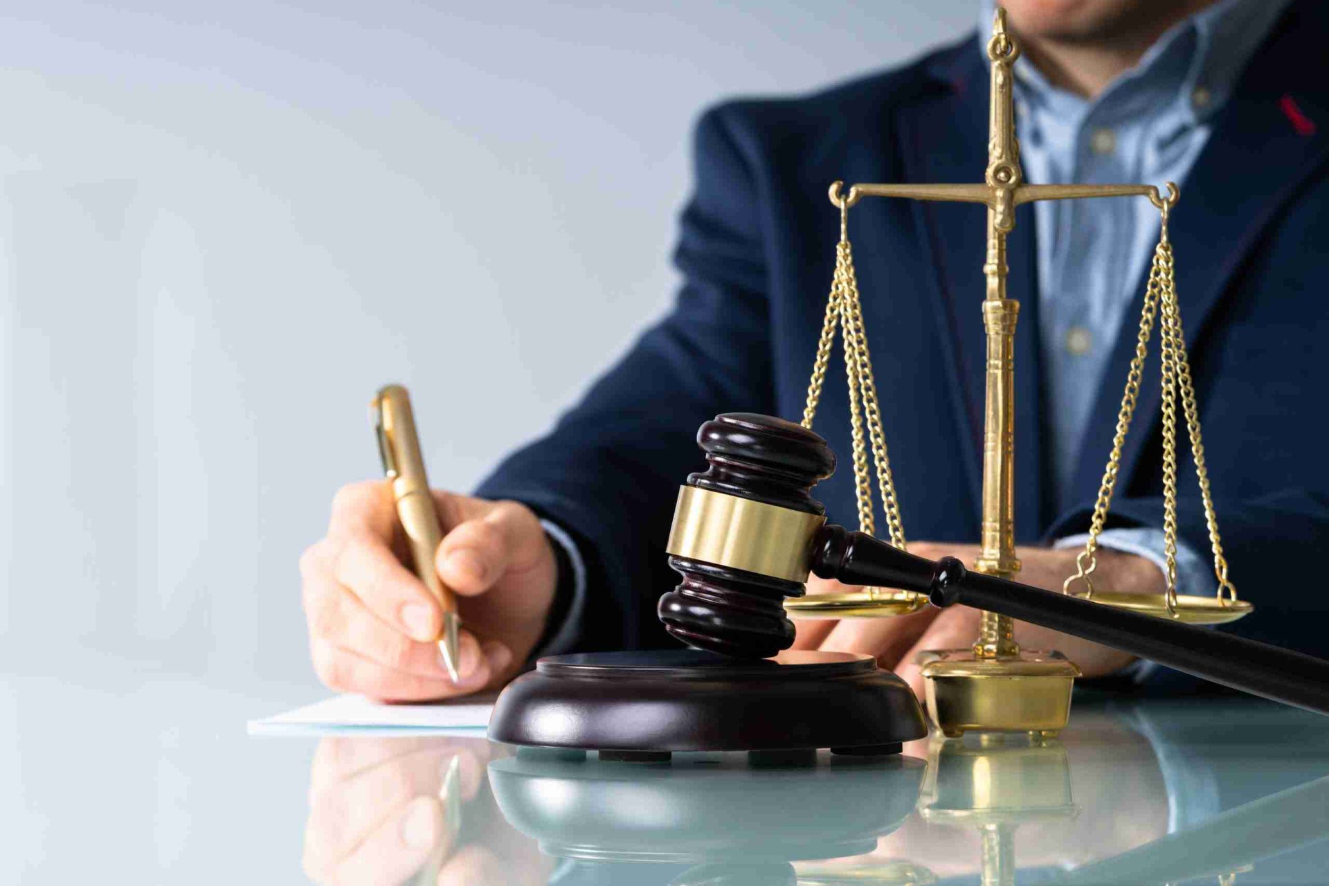 A person in a blue blazer is writing on a piece of paper at a glass table. In front of them, there is a brass balance scale and a judge's gavel resting on its block, symbolizing legal or judicial work.