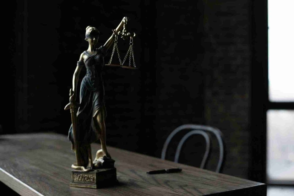 A bronze statuette of Lady Justice holding scales and a sword stands on a wooden table in a dimly lit room. A pen lies nearby, and an empty chair is partially visible in the background against a window.