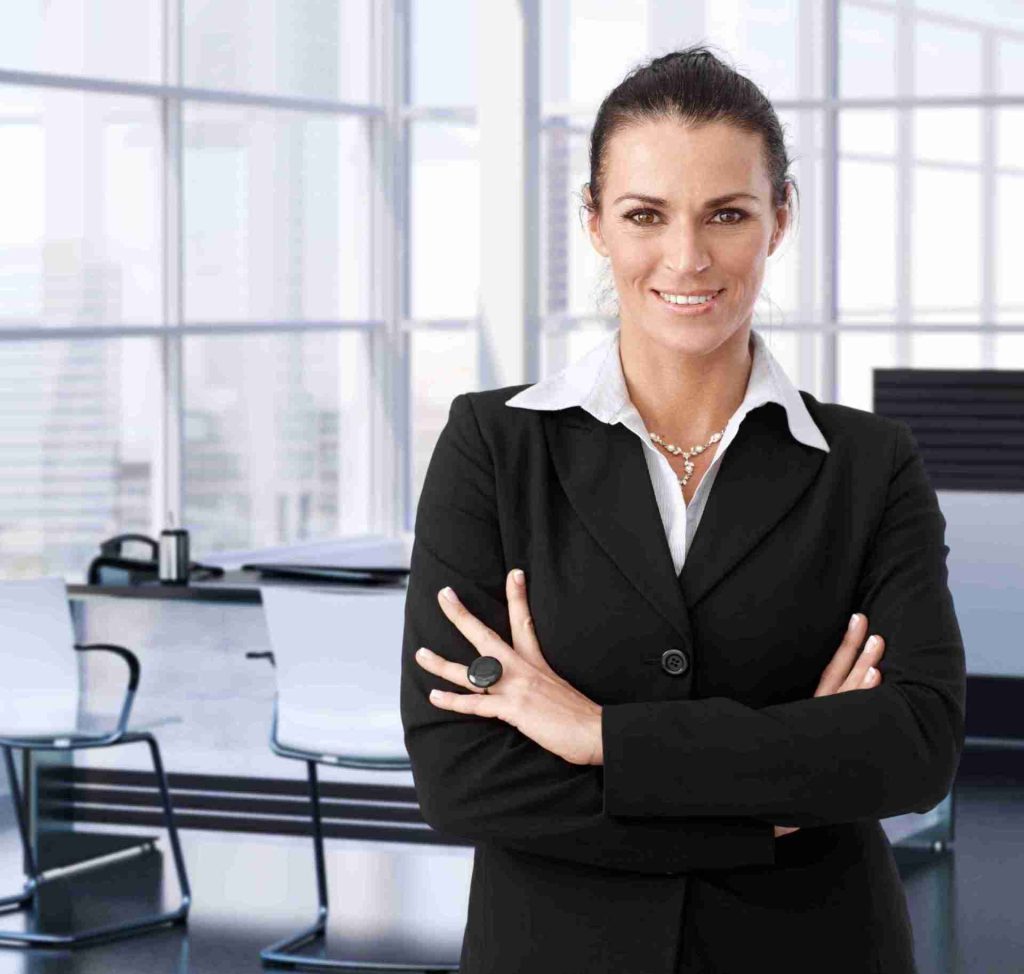 A confident woman with dark hair tied back, wearing a black blazer and white shirt, stands with her arms crossed in a modern office setting. The office has large windows and sleek furniture, including white chairs and a desk with a laptop and coffee cups.