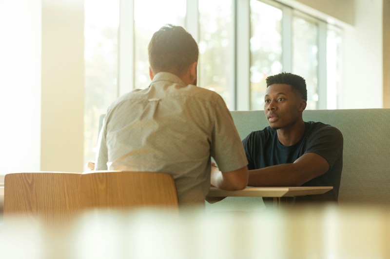 Two men are sitting across from each other at a table in a well-lit room with large windows. One is talking while the other listens attentively. The setting appears casual and relaxed, suggesting a conversation or an informal meeting.