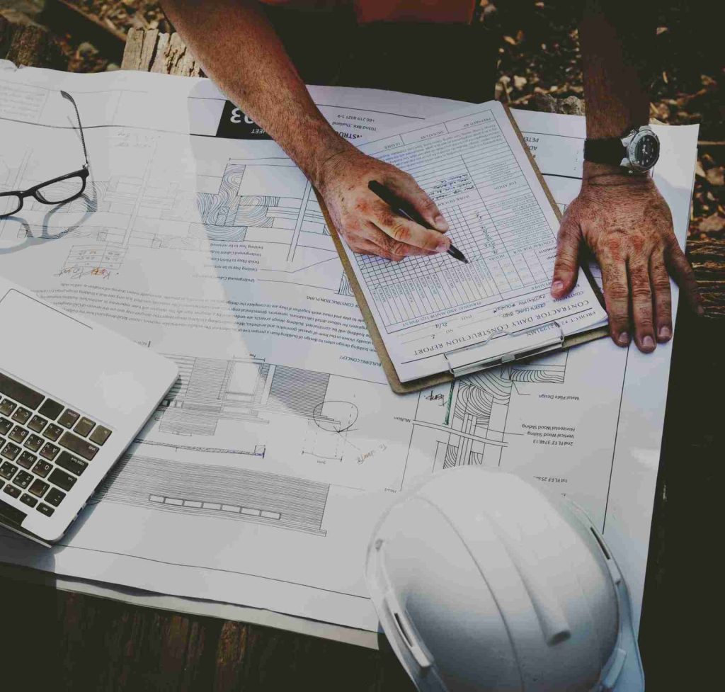 A person is working on a construction site, planning with blueprints and a clipboard. A laptop, glasses, and a white hard hat are on the table. The scene suggests project management and organizing construction tasks.