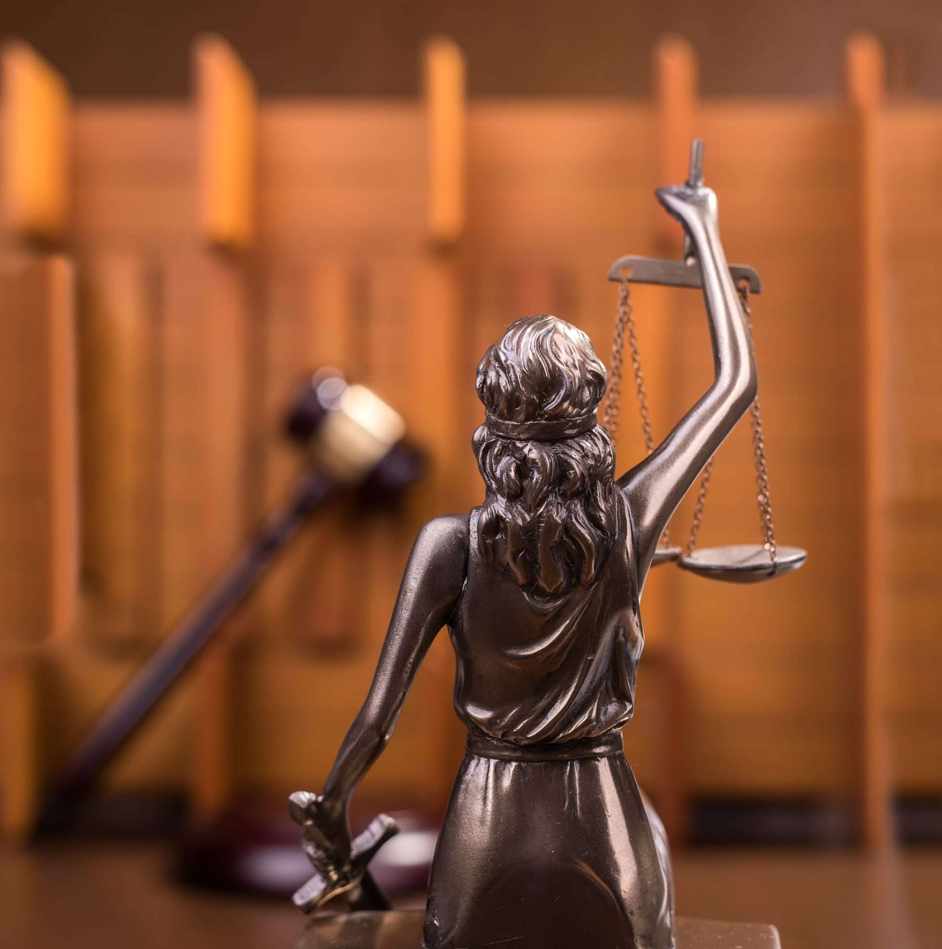 A bronze statue of Lady Justice is prominently displayed, holding scales in her left hand and a sword in her right hand. A blurred gavel and judge's bench are visible in the background, creating a courtroom setting.
