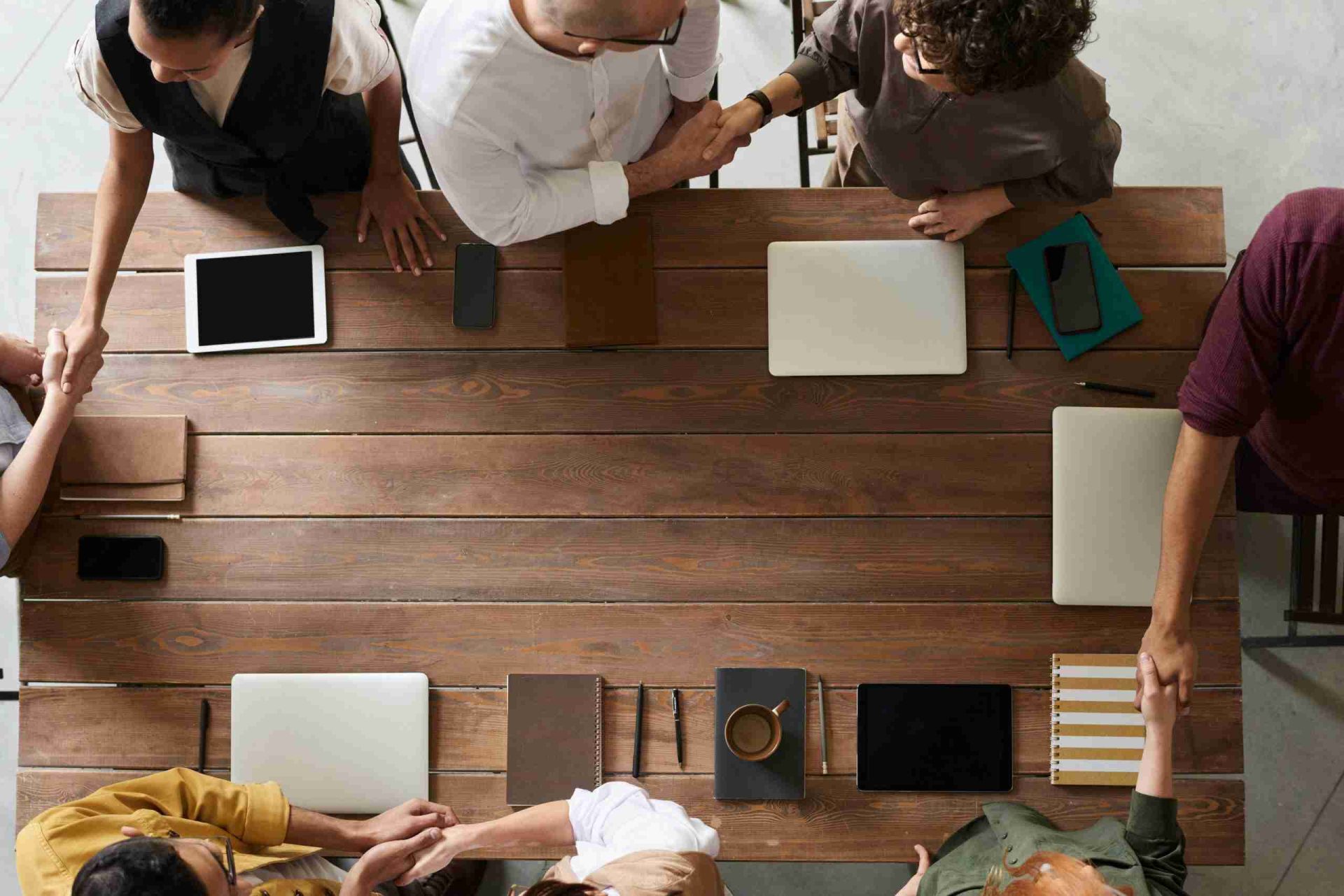 A top-down view of a wooden conference table with various office items such as laptops, tablets, notebooks, phones, and a coffee cup. Six people around the table are engaged in discussions, with some shaking hands, suggesting collaboration or agreement.