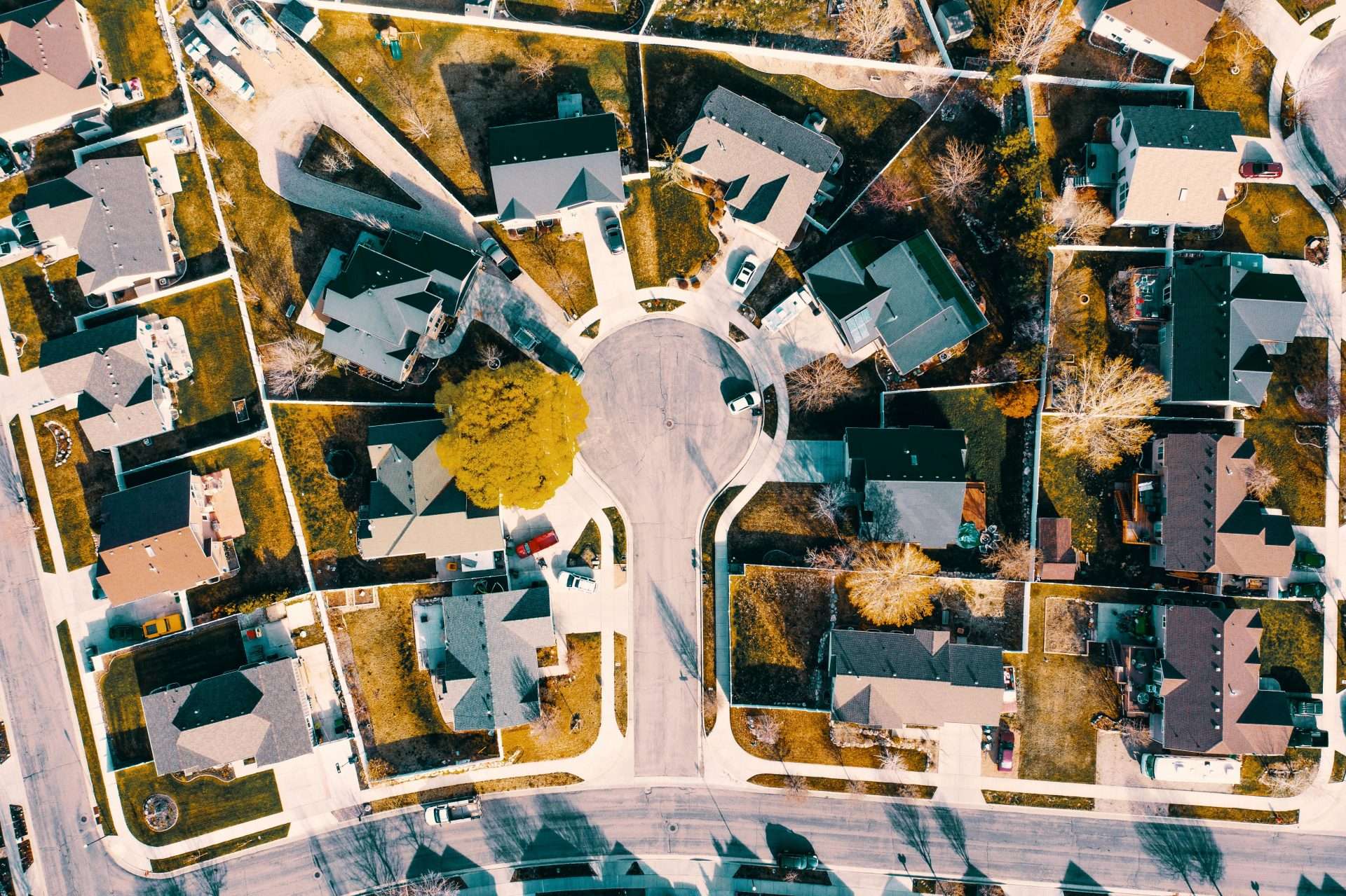 Aerial view of a suburban neighborhood showing closely packed houses with lawns. A central roundabout is visible, with houses arranged around it. Roads, driveways, and a few cars are also seen, along with some trees scattered throughout the area.