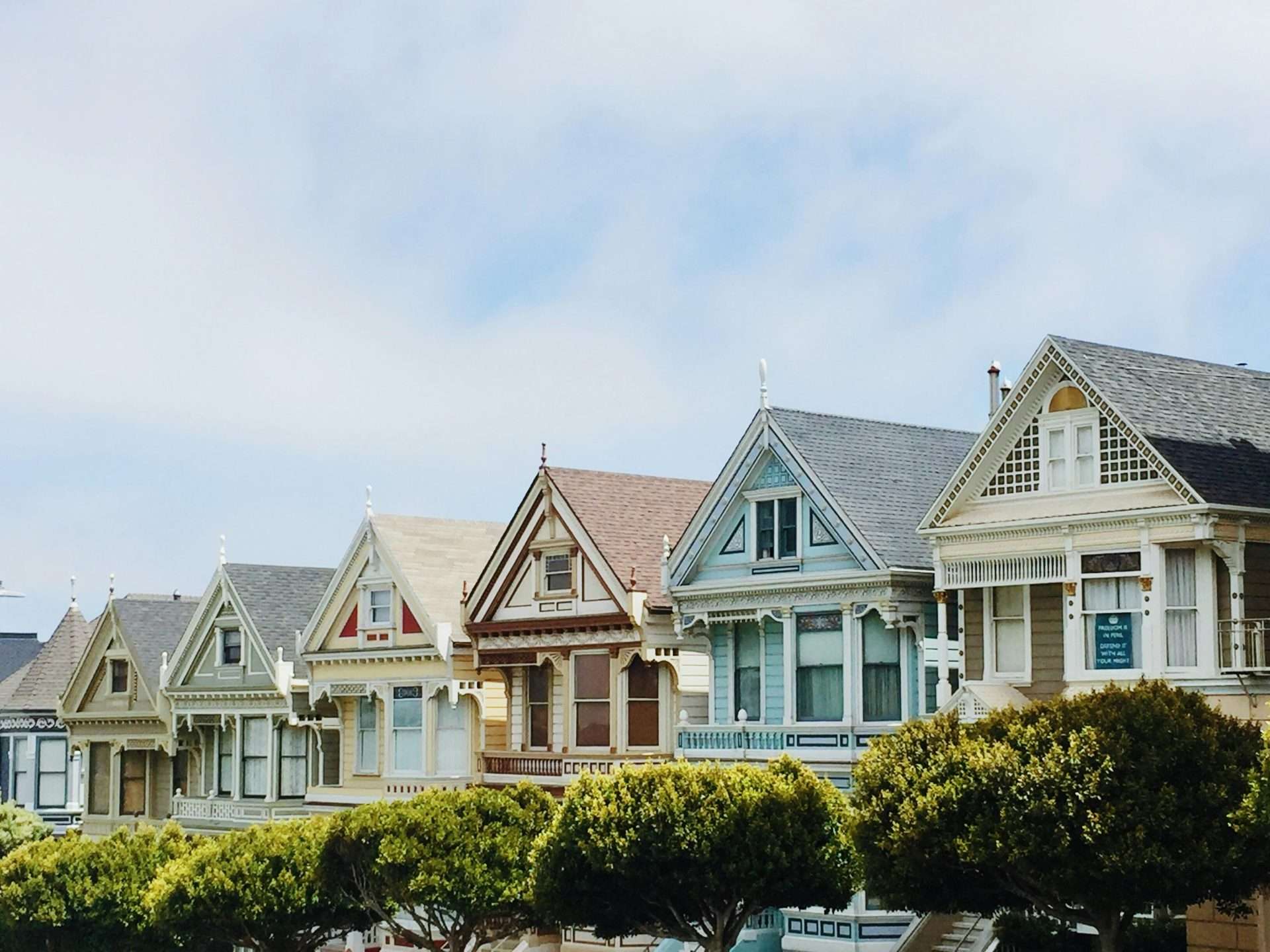 A row of historic Victorian-style houses, known as the Painted Ladies, stands against a blue sky with scattered clouds. Each house features distinct pastel colors and ornate architectural details. Lush green trees line the foreground.
