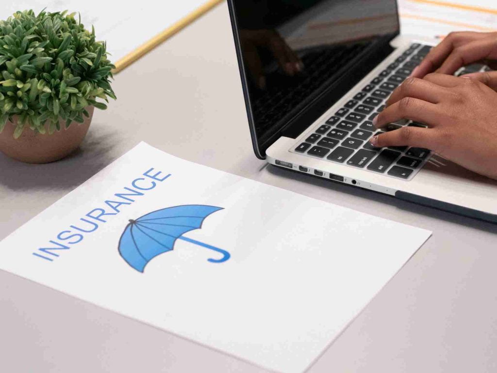A person types on a laptop at a desk. Next to the laptop is a document with an illustration of a blue umbrella and the word "INSURANCE" written in large blue letters. A small potted plant is also visible on the desk.