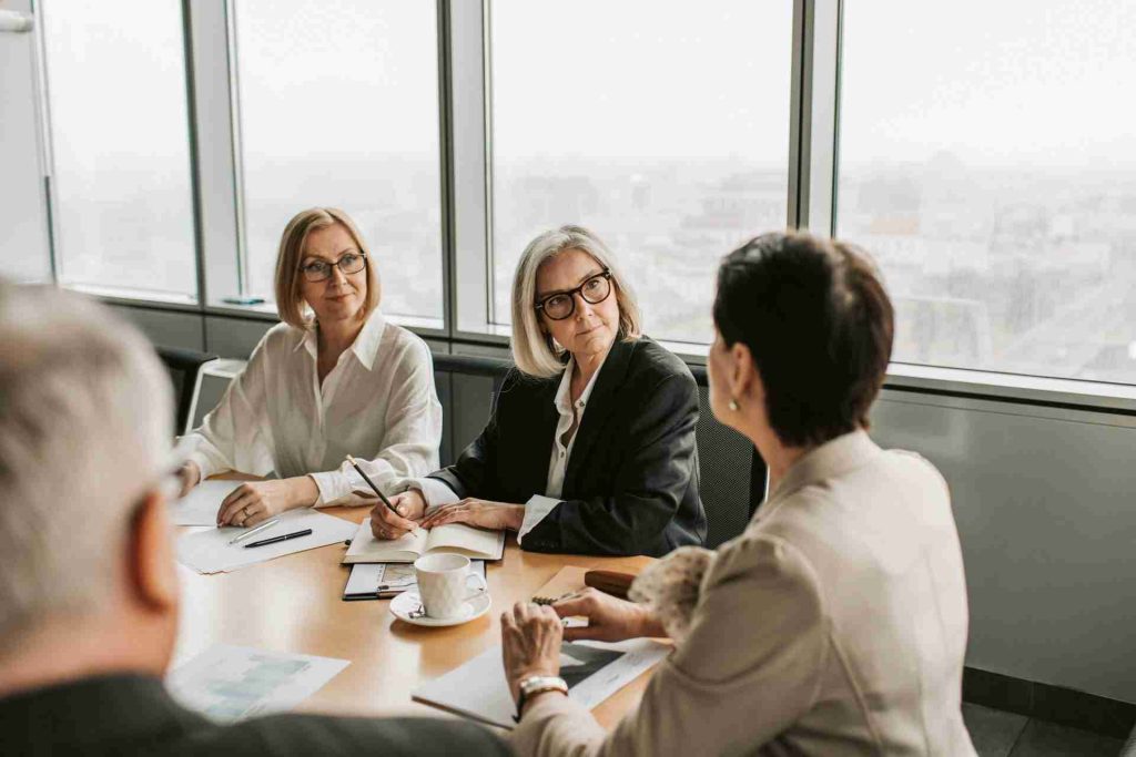 A group of people in professional attire sit around a conference table in a meeting room with large windows. Two women are seen closely listening to another woman who is speaking, while another individual is partially visible. Documents and a coffee cup are on the table.