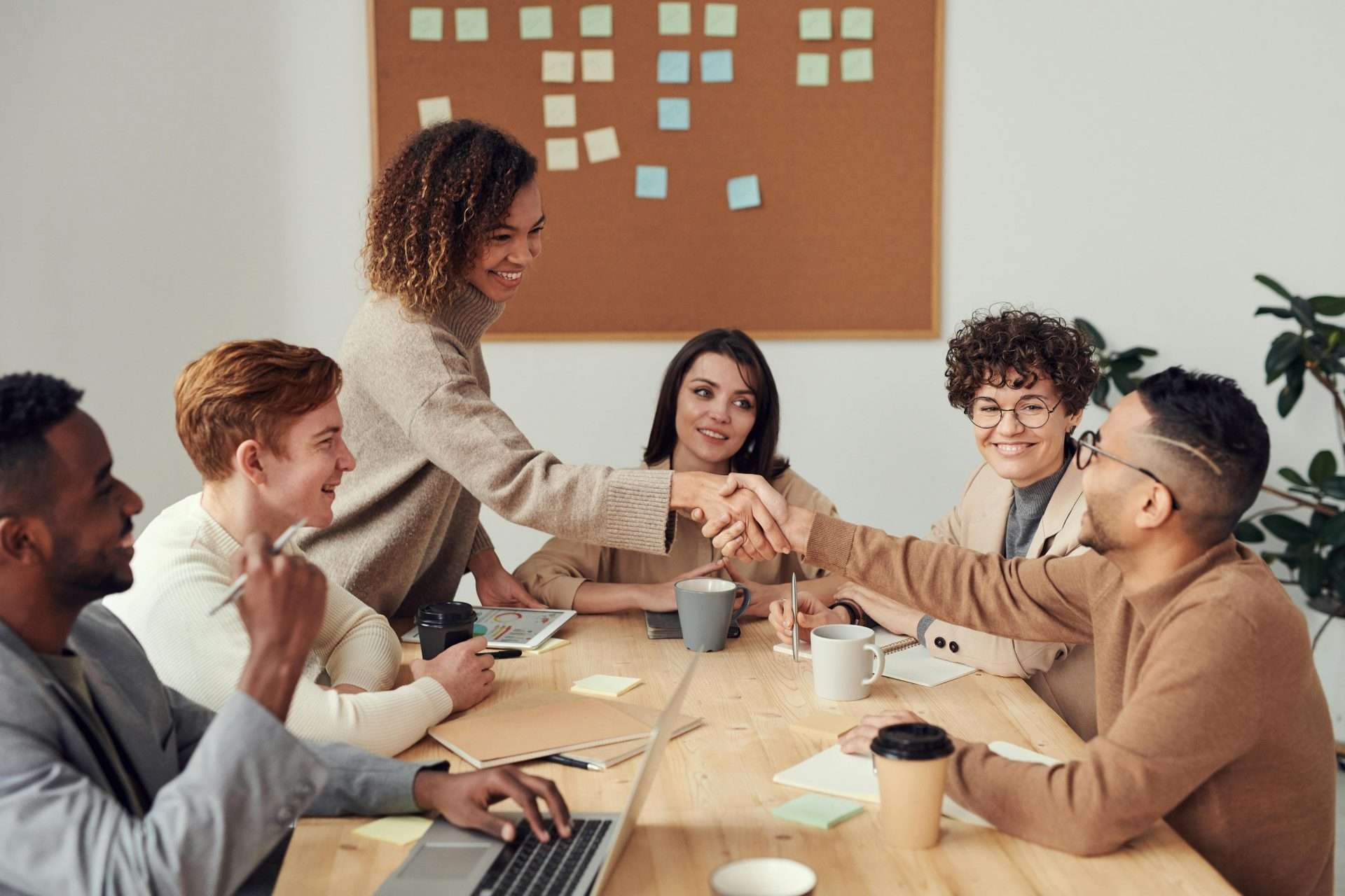 A group of six diverse people sit around a wooden table in an office, engaging in a meeting. One woman stands and shakes hands with a seated man, smiling. Laptops, papers, sticky notes, and coffee cups are on the table. A bulletin board with sticky notes is in the background.