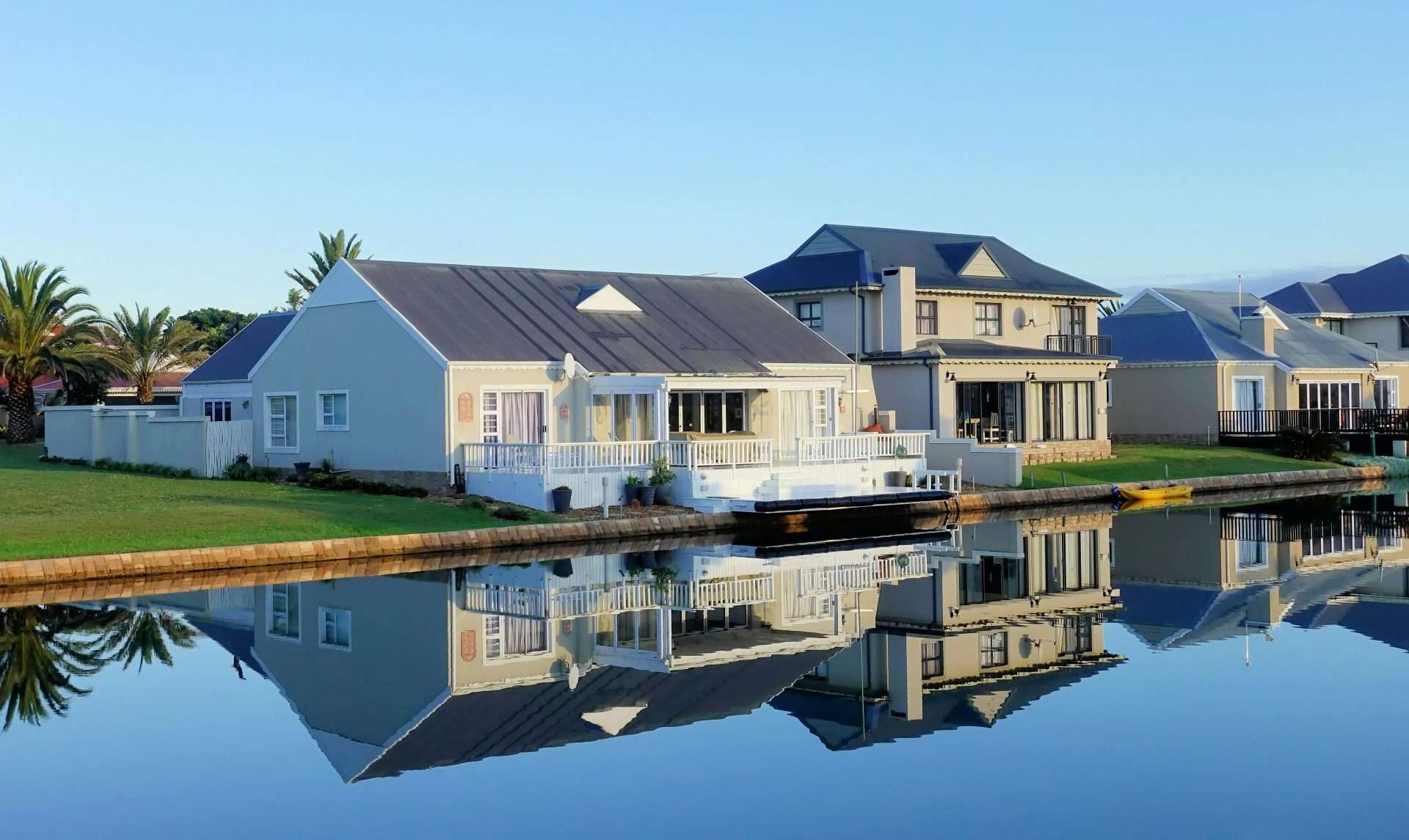 A serene waterfront scene featuring two modern houses with reflective windows and sloped roofs, set along a calm canal. Palm trees and lush lawns surround the homes, with clear blue skies above and perfect reflections on the water’s surface below.