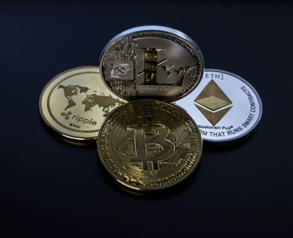 Four cryptocurrency coins are displayed on a black surface. The coins are Ripple (XRP), Litecoin (LTC), Ethereum (ETH), and Bitcoin (BTC), each showing their respective logos and designs with intricate details.