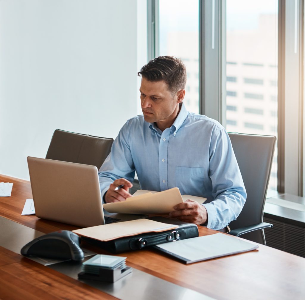 A man in a light blue shirt sits at a wooden conference table, focused on a laptop and holding a folder. The table has a few documents, a pen, and a conference phone. Large windows in the background reveal a blurred cityscape.